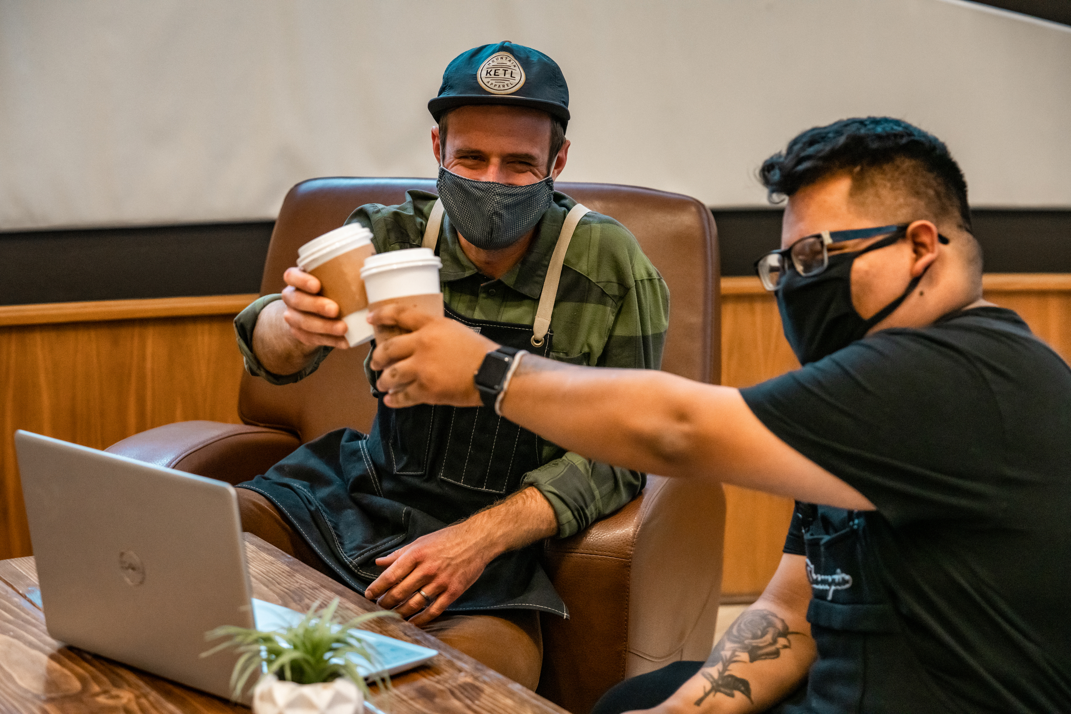Two people are raising their coffee cups together like they are making a toast. They are both wearing face masks while sitting at a table together.