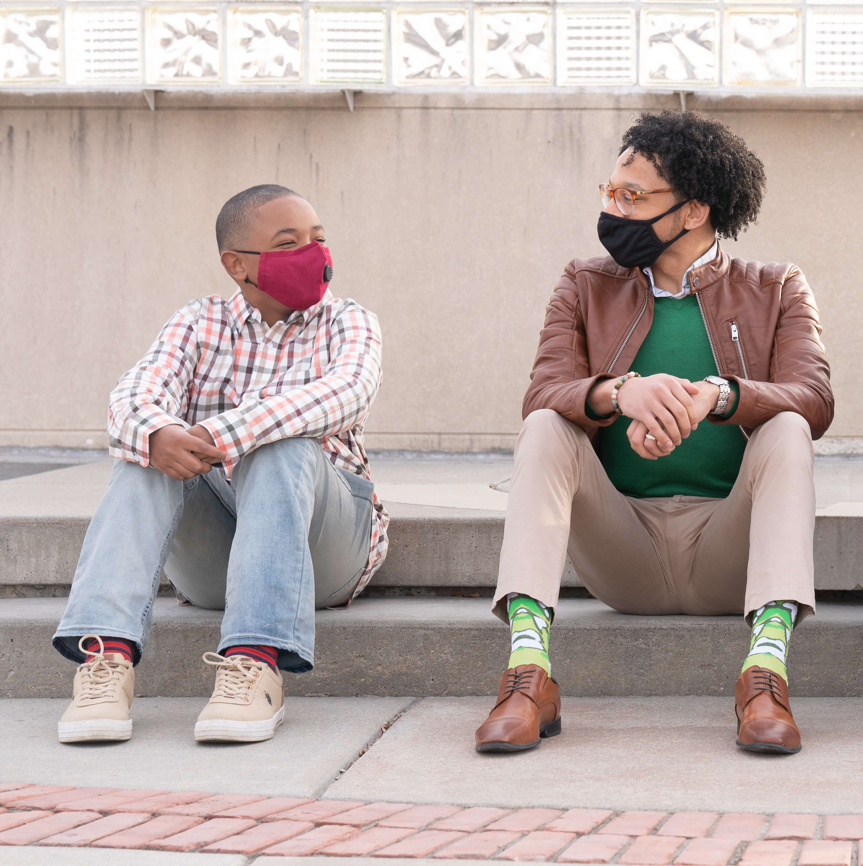 A mentor sitting with a child on a curb and both are masked
