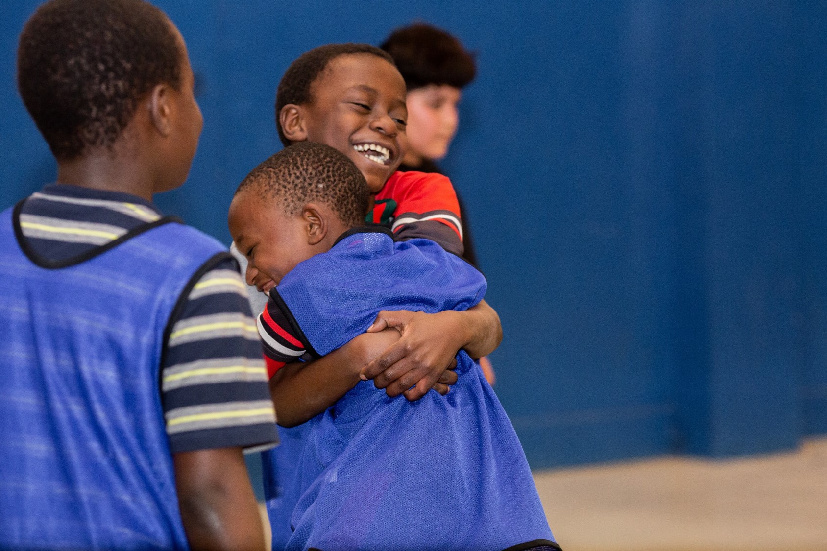 Two young African American boys are grinning and hugging each other. One of them is wearing a blue pinny over his shirt; they are playing a game in a gym setting.