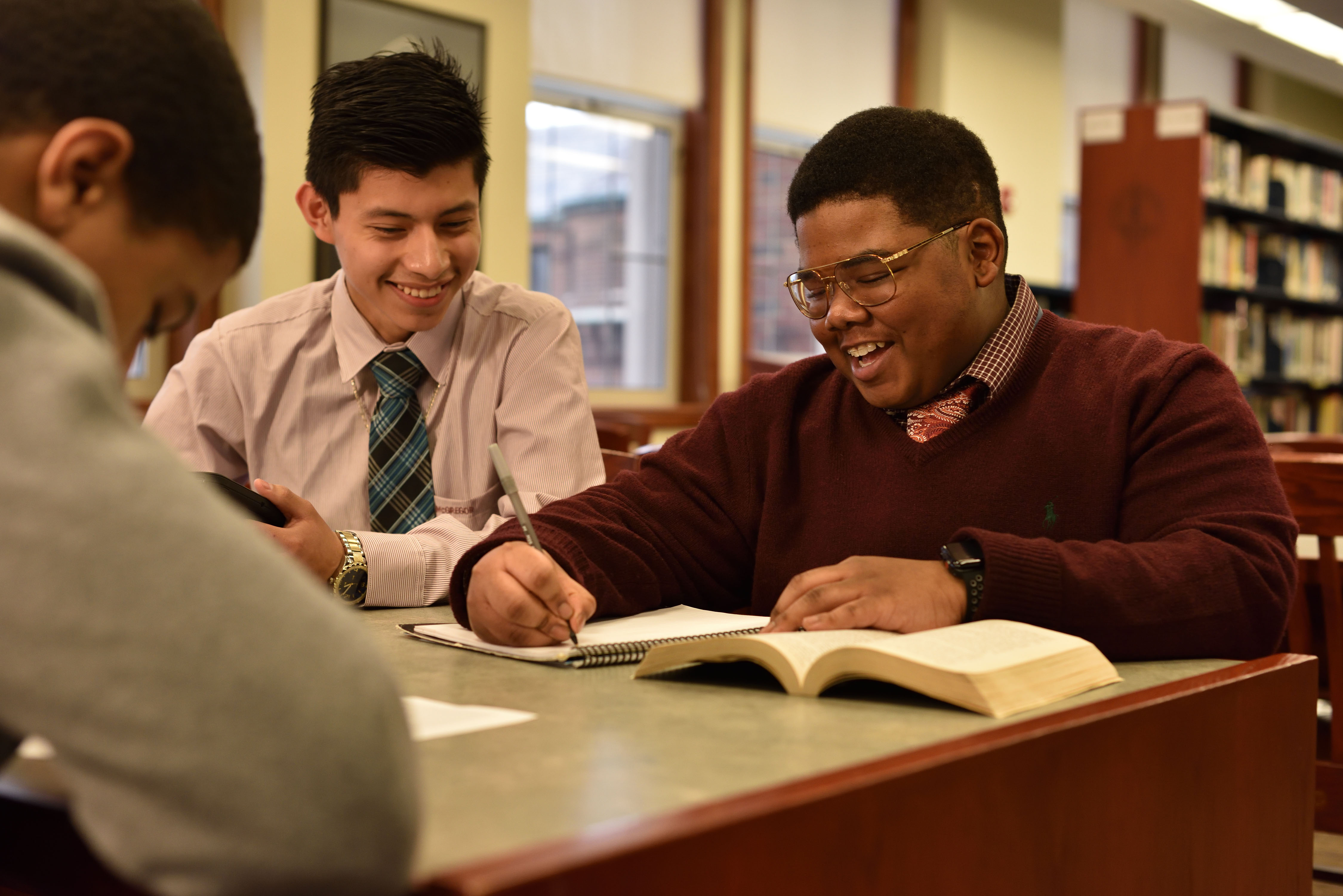 A group of three boys are working at a table together in a library setting. The main subject is a high-school aged African American boy wearing gold glasses and a red sweater. They are all smiling.